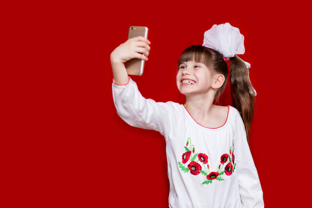 Το χαριτωμένο μικρό κορίτσι σε λευκό τόξα κρατήστε τηλέφωνο και πάρτε την εικόνα σε κόκκινο φόντο. Έννοια επικοινωνίας - Φωτογραφία, εικόνα