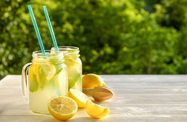 Two mason jar glasses of homemade lemonade, slices of organic ripe lemon, whole and halved, mint, juicer on white wooden table, country side tree foliage background. Close up, top view, copy space. - Фото, зображення