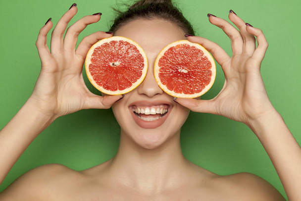 Happy young woman posing with slices of red grapefruit on her face on a green background - 写真・画像