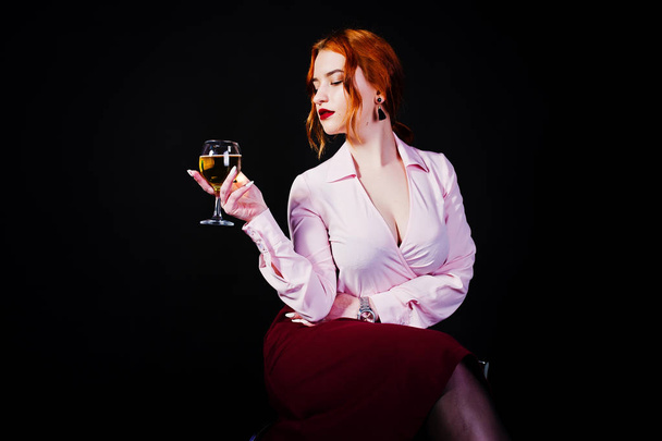 Gorgeous red haired girl in pink blouse and red skirt with glass of wine at hand isolated on black. - Foto, imagen
