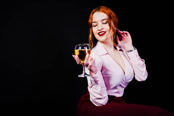 Gorgeous red haired girl in pink blouse and red skirt with glass of wine at hand isolated on black. - Fotó, kép