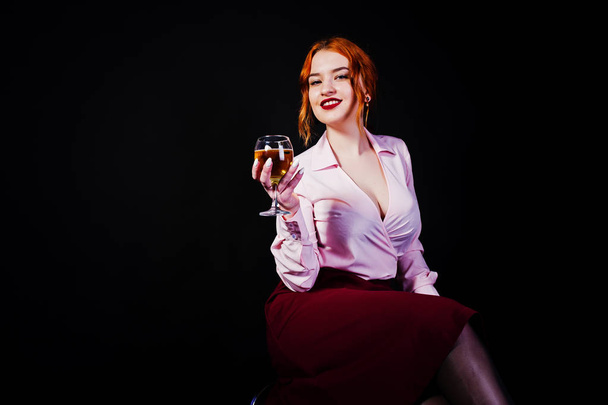 Gorgeous red haired girl in pink blouse and red skirt with glass of wine at hand isolated on black. - Valokuva, kuva