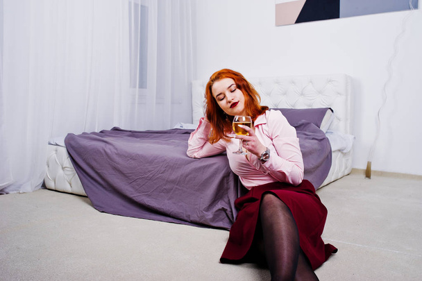 Gorgeous red haired girl in pink blouse and red skirt with glass of wine at hand near bed at room. - Φωτογραφία, εικόνα