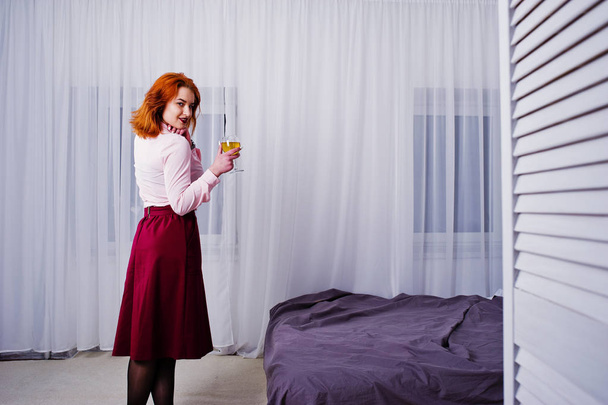 Superbe fille aux cheveux rouges en chemisier rose et jupe rouge avec verre de vin à portée de main près du lit dans la chambre
. - Photo, image