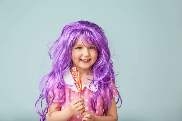 Милая маленькая девочка в фиолетовом парике и с леденцом на цветном фоне
 - Фото, изображение