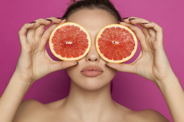 Sexy mladá žena pózuje s plátky červeného grapefruitu na její obličej na růžovém pozadí - Fotografie, Obrázek