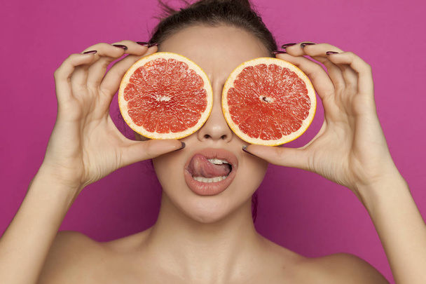 Szexi fiatal nő pózol a szelet az arcán, egy rózsaszín háttér vörös grapefruit - Fotó, kép