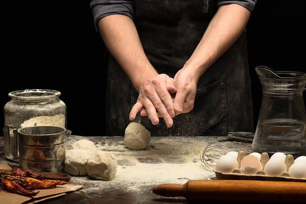 Cooks hands are rolling dough on wooden table with white wheat flour. Black background - Photo, Image