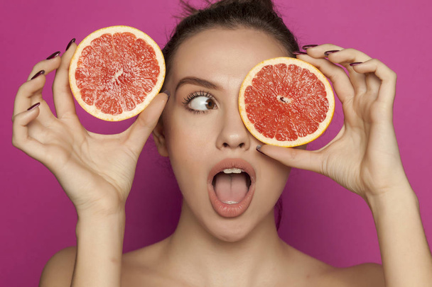 Young surprised woman posing with slices of red grapefruit on her face on a pink background - Foto, afbeelding