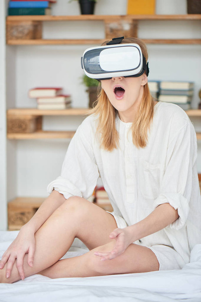 astonished woman with opened mouth using virtual reality headset while sitting on bed at home    - Φωτογραφία, εικόνα