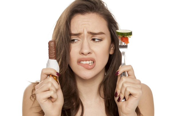 Jovem mulher bonita é tentada por chocolate e vegetais em um fundo branco
 - Foto, Imagem