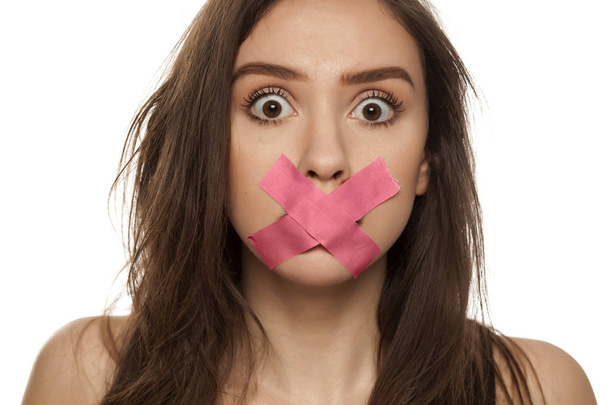 Mladá žena překvapená s lepící páskou přes ústa na bílém pozadí - Fotografie, Obrázek