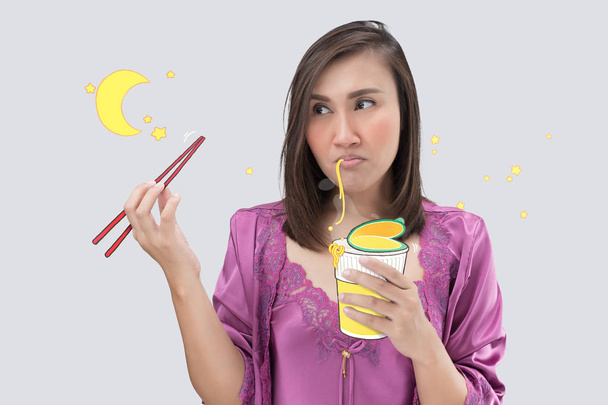 Азиатка в атласной фиолетовой ночной рубашке, ест по ночам чашку лапши. Женщина голодна по ночам на сером фоне. Лапша быстрого приготовления
. - Фото, изображение