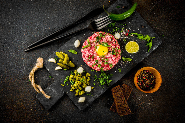 French cuisine, beef steak tartare with raw quail egg yolk, pickled cucumber and onions, fresh herbs, bread, dark rusty background copy space - Photo, Image