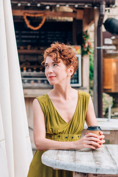 A beautiful redhead woman in a green dress stands, holds coffee in her hands and looks away with a smile. Summer, relax, vacation concept - Foto, Imagem