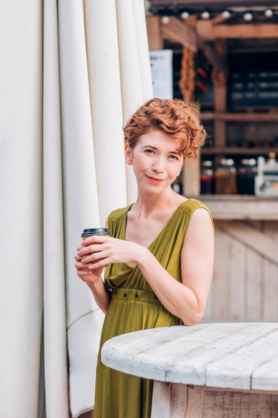 Uma mulher bonita em um café de verão com café em suas mãos olha para a câmera com um sorriso
 - Foto, Imagem