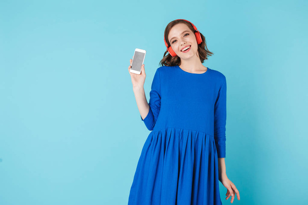 Portrait de jeune femme souriante en robe bleue et écouteurs debout avec téléphone portable et regardant joyeusement à la caméra sur fond rose
 - Photo, image