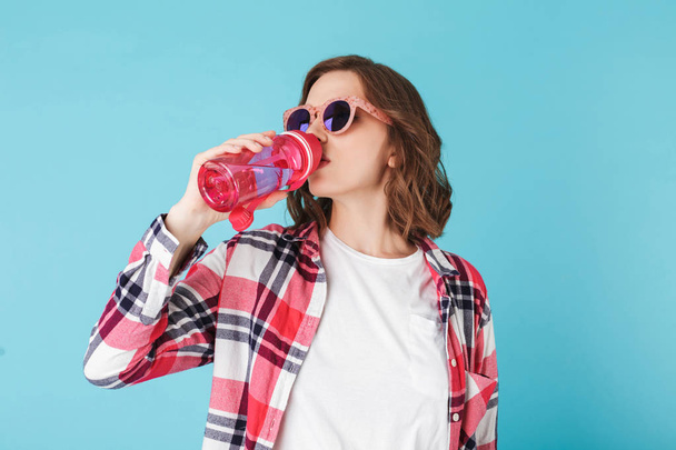 Krásná žena v sluneční brýle stojí s růžové sportovní láhev a pití vody nad barevné pozadí - Fotografie, Obrázek