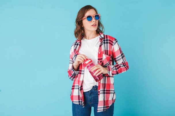 Beautiful lady in sunglasses standing with pink sport bottle over blue background Photo of nice hipster girl in plaid shirt and jeans - Photo, Image