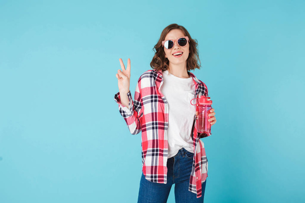 Beautiful smiling lady in sunglasses standing with pink sport bottle over pink background Photo of pretty girl in plaid shirt and jeans showing peace and victory sign gesture - Photo, Image