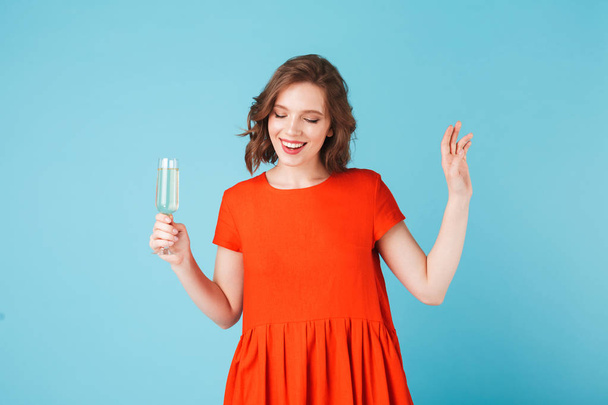 Beautiful lady in red dress standing with champagne and happily looking aside over colorful background - Photo, Image