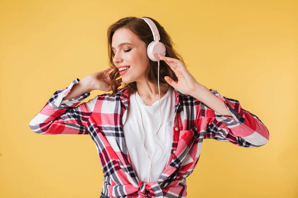 Portrait of young smiling lady in colorful shirt standing and listening music in headphones on over yellow background - Фото, изображение