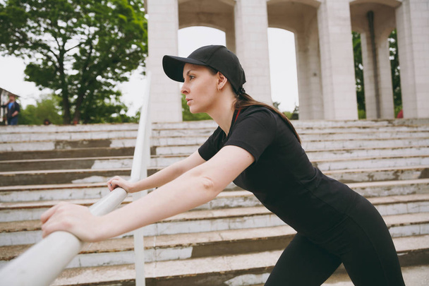 Young concentrated athletic beautiful brunette girl in black uniform and cap doing sport stretching exercises, warm-up before running in city park outdoors. Fitness, healthy lifestyle concept - Фото, зображення