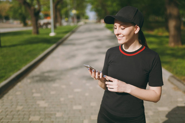 Αθλητική χαμογελώντας όμορφη μελαχρινή κοπέλα με μαύρη στολή και καπάκι χρήση κινητού τηλεφώνου κατά τη διάρκεια της κατάρτισης, ψάχνει στο smartphone, στέκεται στο πάρκο της πόλης σε εξωτερικούς χώρους. Γυμναστήριο, έννοια του υγιεινού τρόπου ζωής - Φωτογραφία, εικόνα