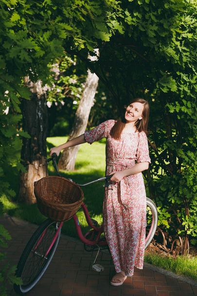 Portrait of trendy young woman in long pink floral dress stop to riding under oak arch on bike with basket for purchases food or flowers outdoors, cute female recreation time in spring or summer park - Фото, изображение