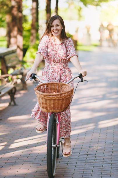 Porträt einer trendigen jungen Frau im langen rosafarbenen floralen Kleid, die auf einem Vintage-Fahrrad mit Korb für Einkäufe, Lebensmittel oder Blumen im Freien reitet, wunderschöne weibliche Freizeit im Frühling oder Sommer Park - Foto, Bild
