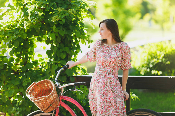 Portret van trendy jonge vrouw in lange roze bloemen jurk stoppen te rijden op vintage fiets met mand voor aankopen, voedsel of bloemen in openlucht, prachtige vrouwelijke recreatie tijd in de lente of zomer park - Foto, afbeelding