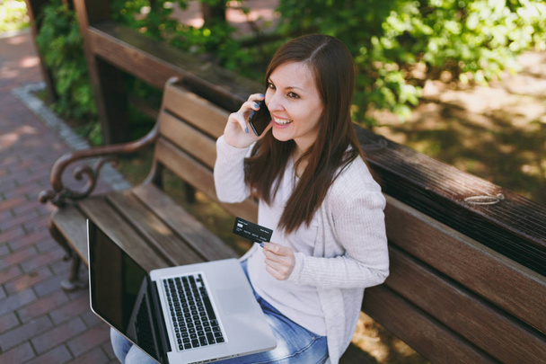 Female holding credit card, talking on mobile phone, having good pleasure conversation. Woman sitting on bench working on modern laptop in street outdoors. Mobile Office. Freelance business concept - Photo, Image