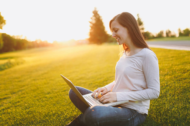 カジュアルな服装の若い学生の女性。サンシャイン芝生を屋外で緑の草の上の都市公園におけるノート パソコン pc コンピューターで作業して地面、草の上に座っている女性。モバイル オフィス。フリーランスのビジネス コンセプト - 写真・画像