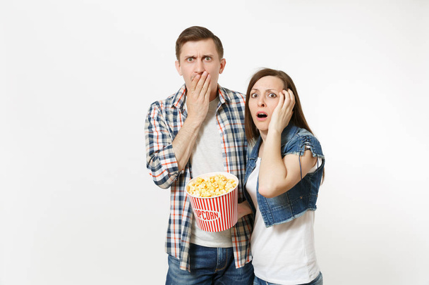 Young shocked scared couple, woman and man watching movie film on date, holding bucket of popcorn, clinging to face and covering mouth isolated on white background. Emotions in cinema concept - 写真・画像