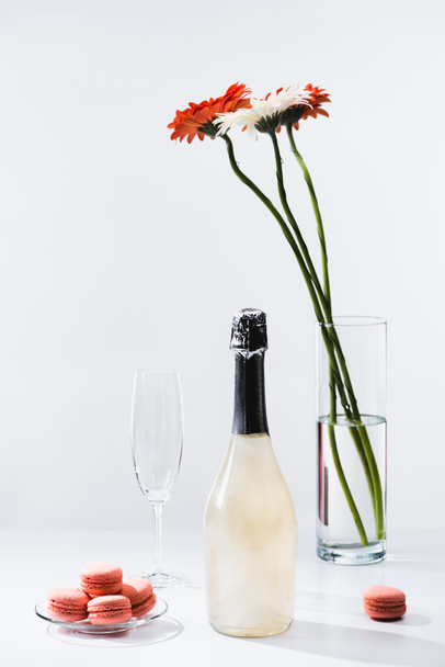 close up view of macarons, empty glass, bottle of champagne and bouquet of gerbera flowers on grey backdrop - Fotoğraf, Görsel