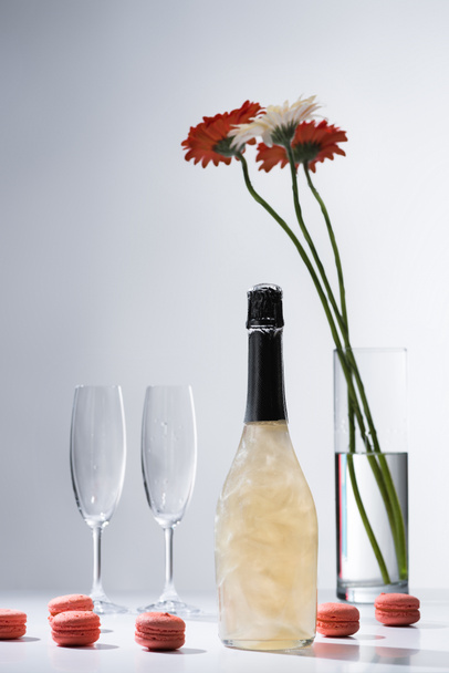 close up view of macarons, empty glasses, bottle of champagne and bouquet of gerbera flowers on grey backdrop - Photo, Image