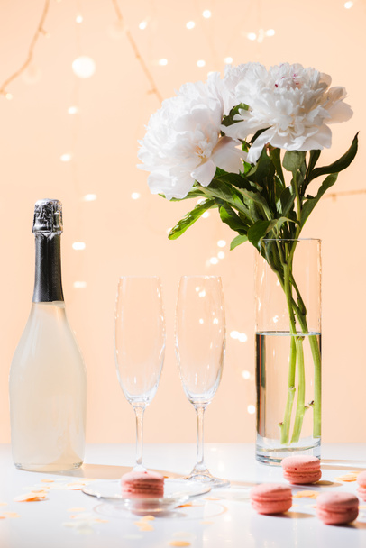 Close-up van fles champagne, lege glazen, macarons en boeket van bloemen van de pioenroos in vaas op roze achtergrond - Foto, afbeelding