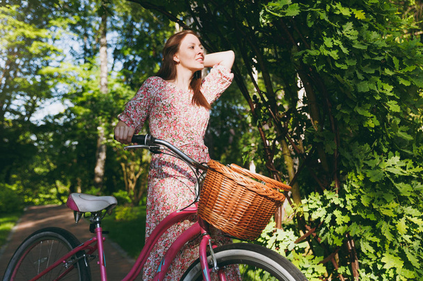 Muotokuva trendikäs nuori nainen pitkä vaaleanpunainen kukka mekko pysähtyy ratsastus tammen kaaren pyörällä korilla ostaa ruokaa tai kukkia ulkona, söpö naisten virkistysaika keväällä tai kesällä puistossa
 - Valokuva, kuva