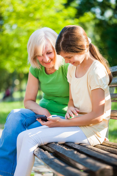 Abuela y nieta están sentadas en el parque y el uso de teléfono móvil
. - Foto, imagen
