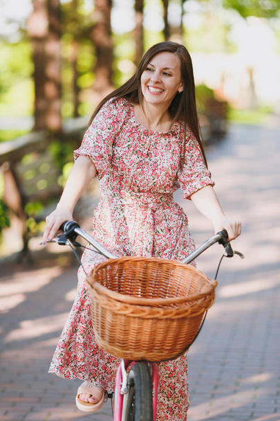 Retrato de mujer joven de moda en vestido largo floral rosa cabalgando en callejón en bicicleta vintage con cesta para compras, comida o flores al aire libre, magnífico tiempo de recreación femenina en el parque de primavera o verano
 - Foto, imagen