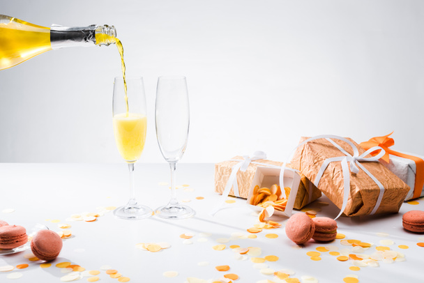close up view of pouring yellow champagne into glasses process, macarons and arranged gifts on grey backdrop - Фото, изображение