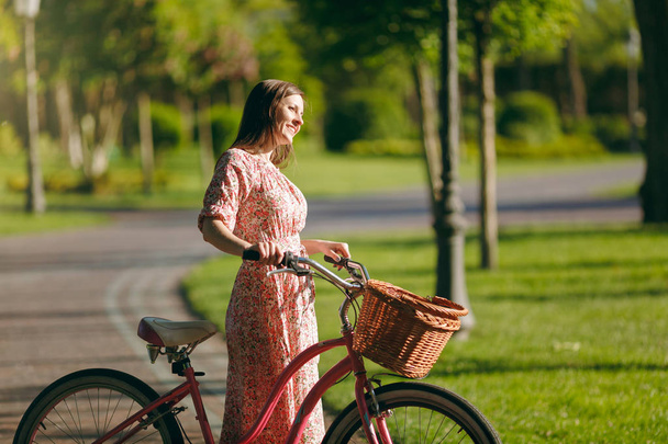 Портрет модної молодої жінки в довгій рожевій квітковій сукні зупиняється, щоб кататися на старовинному велосипеді з кошиком для покупок, їжі або квітів на відкритому повітрі, чудовий жіночий час відпочинку навесні або в літньому парку
 - Фото, зображення