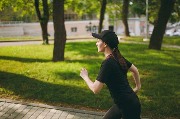 Young concentrated athletic beautiful brunette girl in black uniform and cap training doing sport exercises running looking straight on path in city park outdoors. Fitness, healthy lifestyle concept - Photo, Image