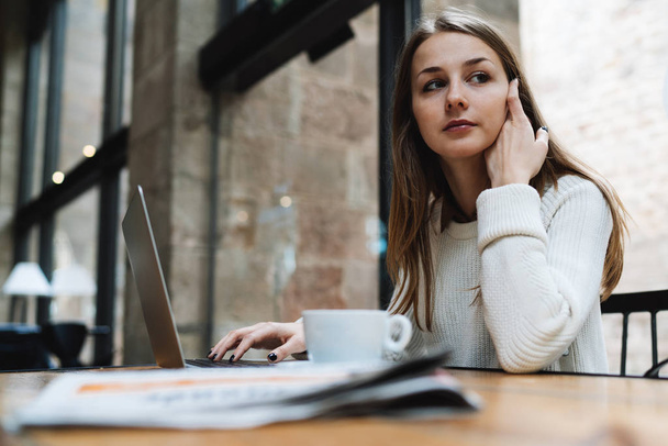Jeune femme regarde de côté tout en étant assis dans un café avec ordinateur portable et une tasse de café sur une table
  - Photo, image