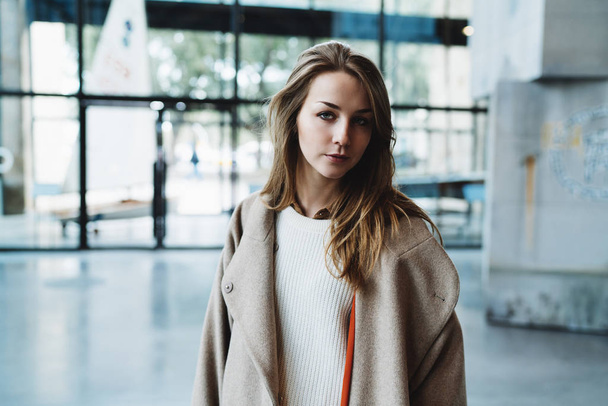 Pensive caucasian female with long blonde hair wearing a coat and a sweater looking at the camera  - Foto, Imagen