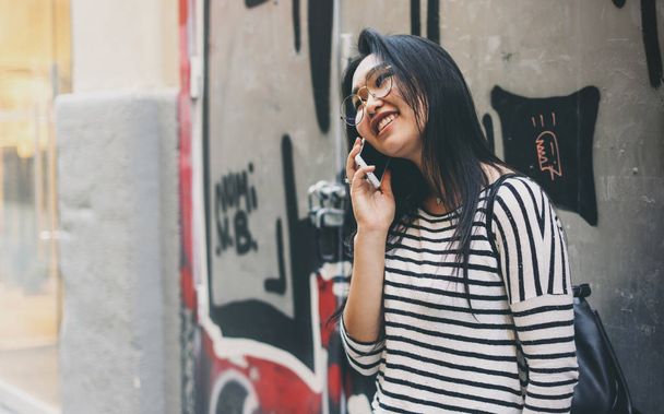落書きの壁背景に立っている間携帯電話で話している若い魅力的なアジア流行に敏感な女性  - 写真・画像