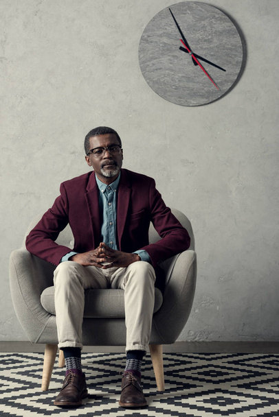 thoughtful african american man sitting in armchair with clock on wall   - Фото, зображення