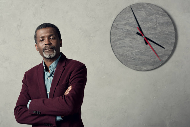 confiado hombre de negocios afroamericano con brazos cruzados de pie cerca de la pared con gran reloj
  - Foto, Imagen