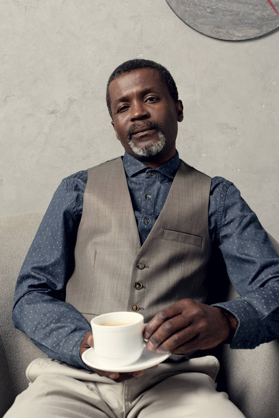 confident stylish african american man sitting in armchair with cup of coffee - Fotografie, Obrázek
