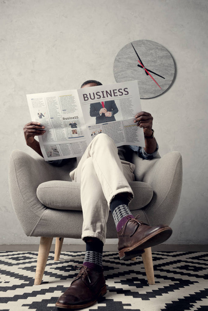 壁の時計を肘掛け椅子に坐っている間ビジネス新聞を読んでスタイリッシュなアフリカ系アメリカ人 - 写真・画像